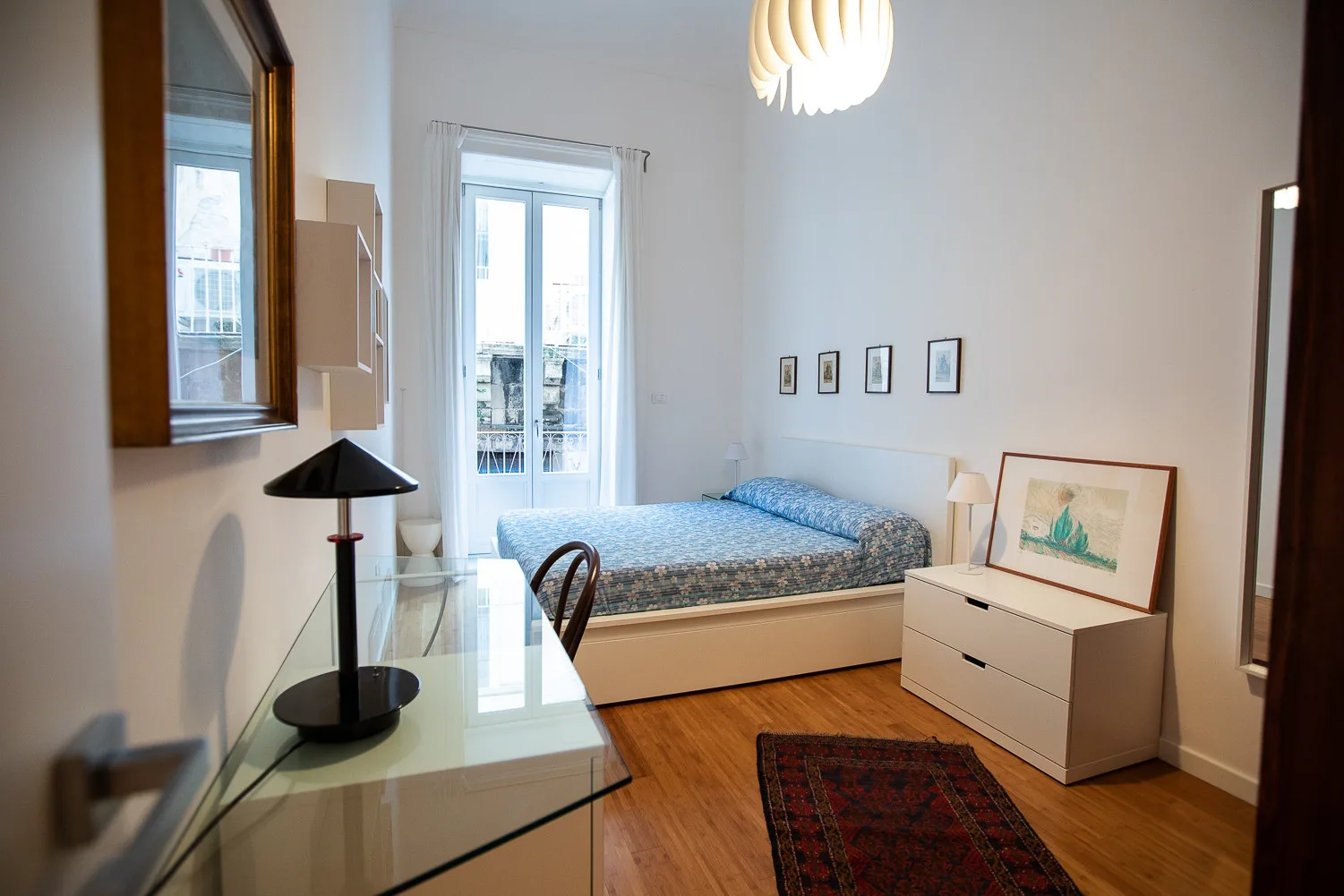 Un dormitorio con una cama, un escritorio de vidrio y ventanas altas.
