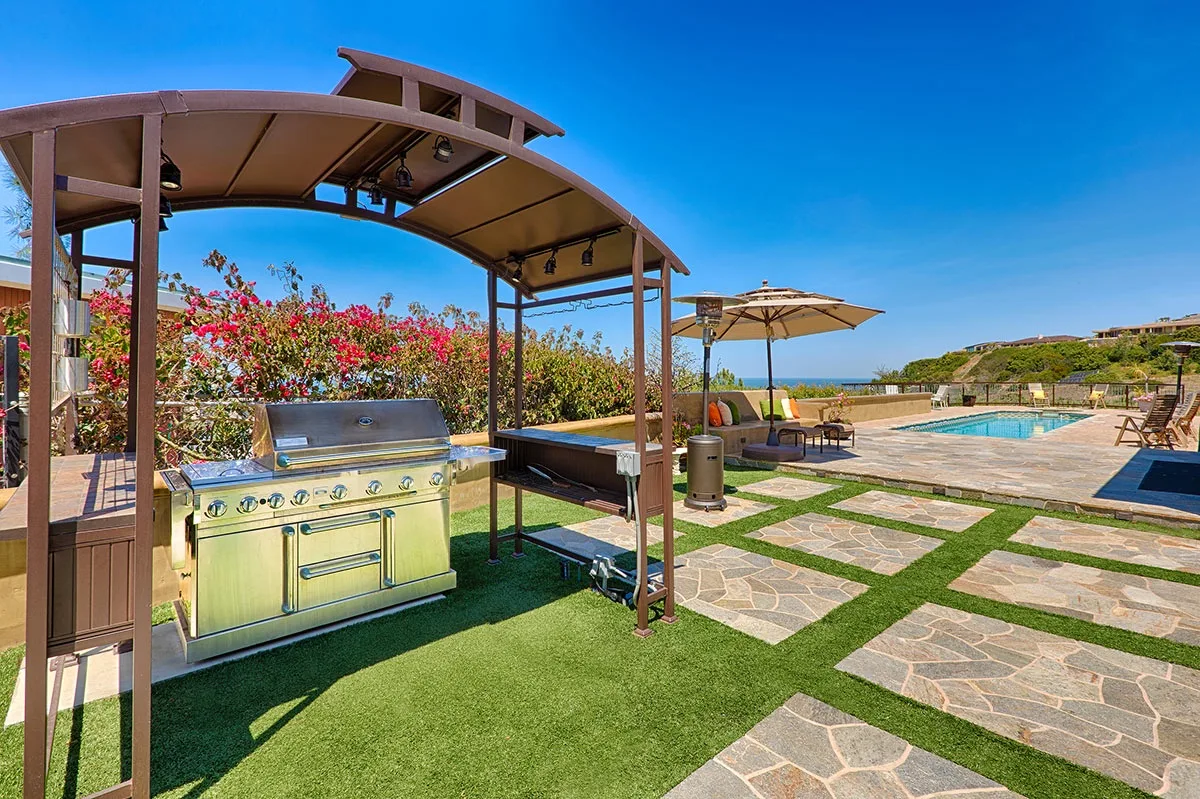 An outdoor kitchen beneath a pergola overlooks a patio and swimming pool.