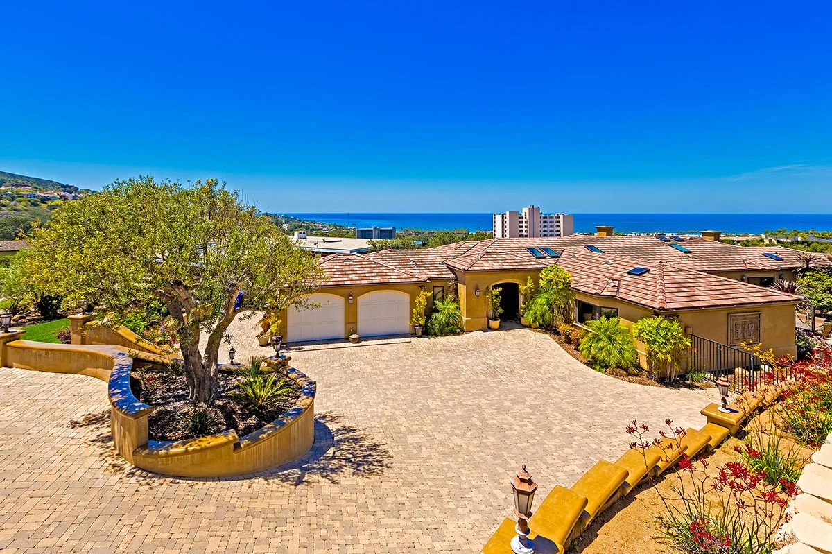 A paved courtyard in front of a low building, with the sea visible in the distance.