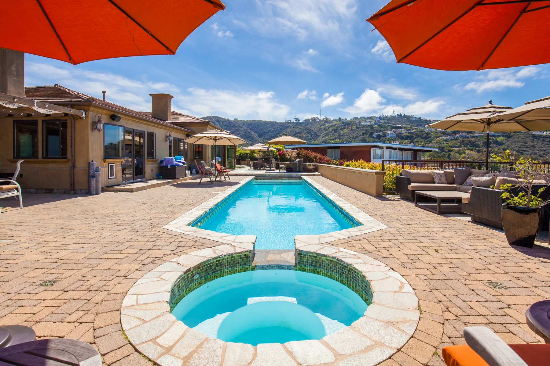 An outdoor pool and spa sit on a sunny patio with seating and umbrellas.