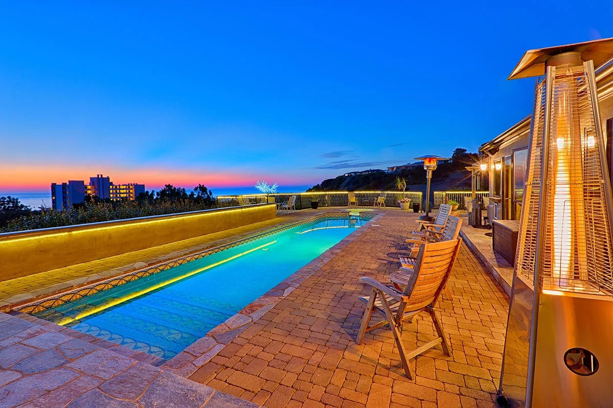 An outdoor pool and surrounding patio seating overlook the coast at sunset.