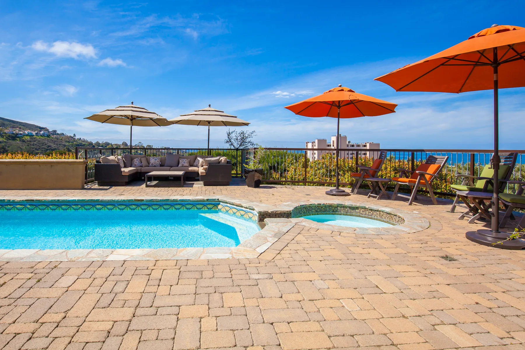 An outdoor pool and hot tub sit beside patio seating and bright umbrellas.