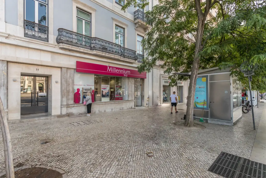 Una calle peatonal pavimentada con una sucursal bancaria, un árbol y un quiosco acristalado.
