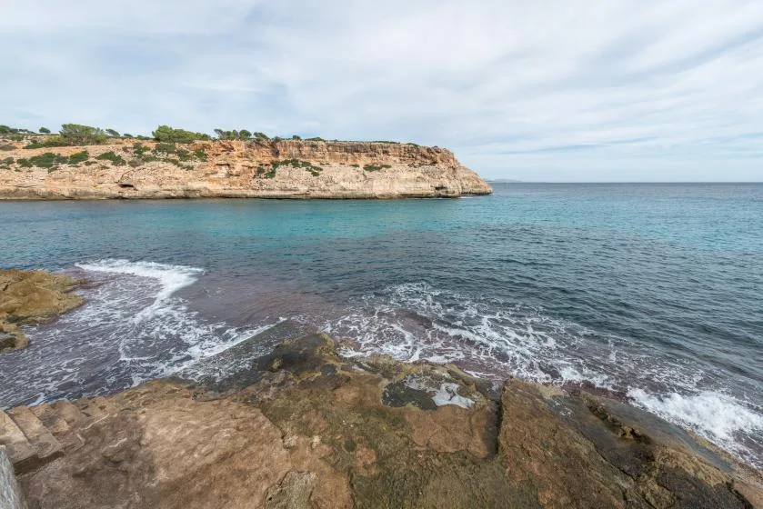 A rocky shoreline meets calm blue water below a low coastal cliff.