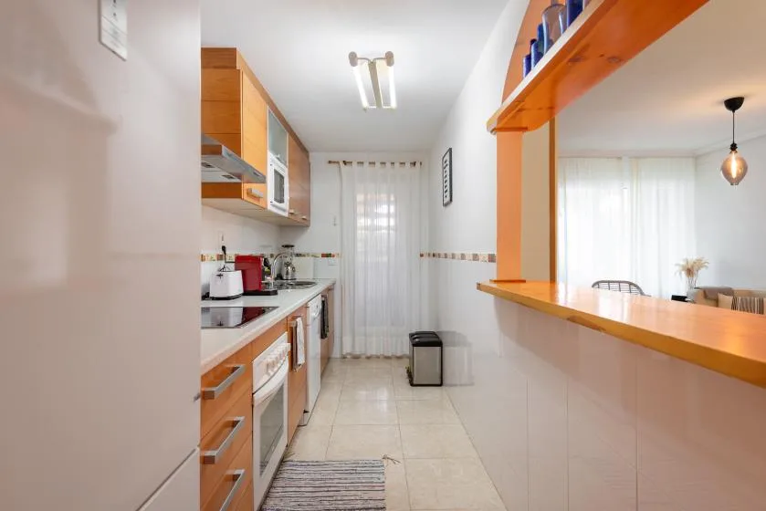A narrow kitchen with wooden cabinets, a countertop, and a curtained window.