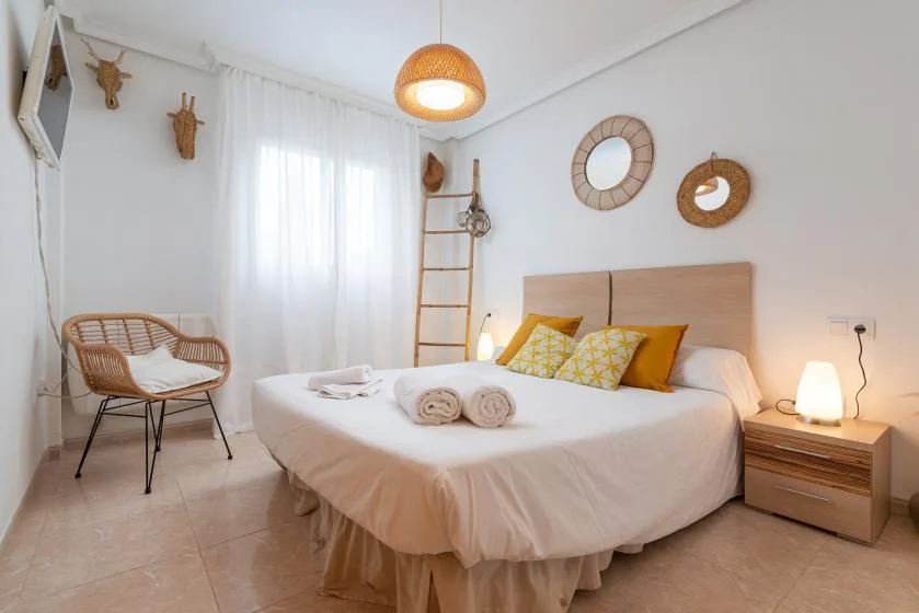 A bright bedroom with a neatly made bed and woven chair beside a curtained window.