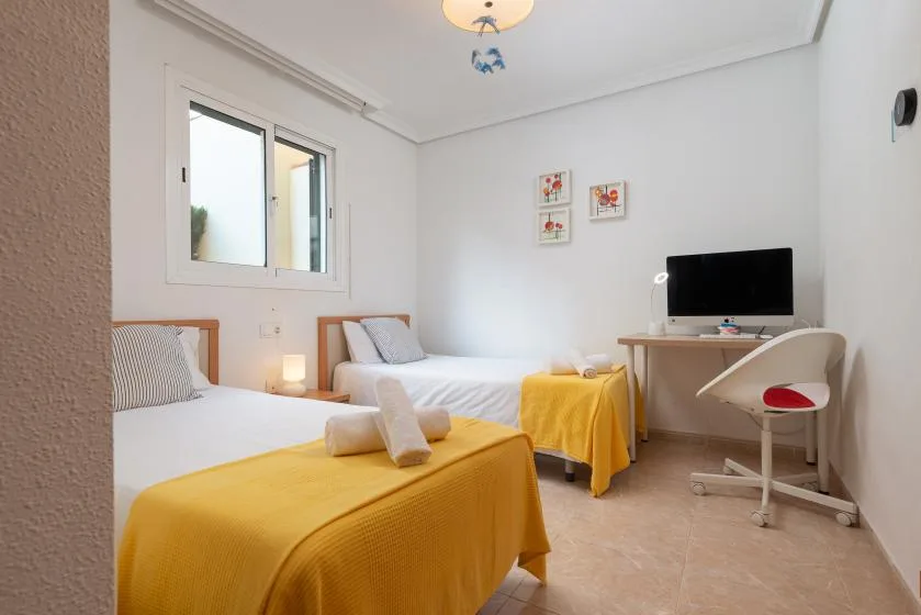A bright bedroom with two beds, yellow bedspreads, and a desk with a monitor.