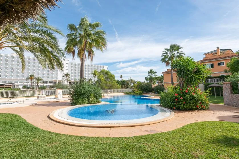 A landscaped outdoor pool area with palm trees, greenery, and surrounding buildings.
