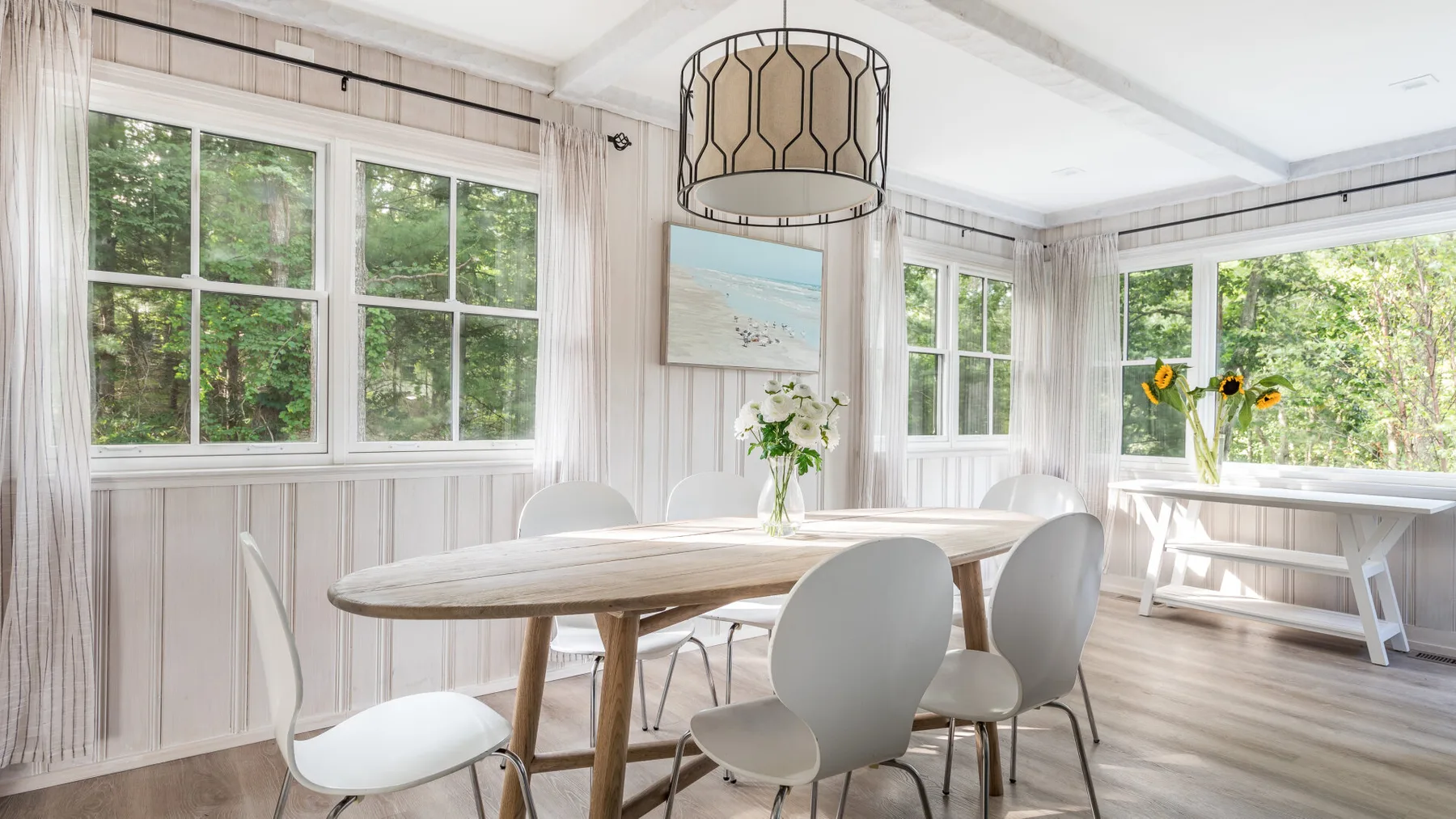Un comedor luminoso con una mesa de madera, asientos blancos y grandes ventanas.