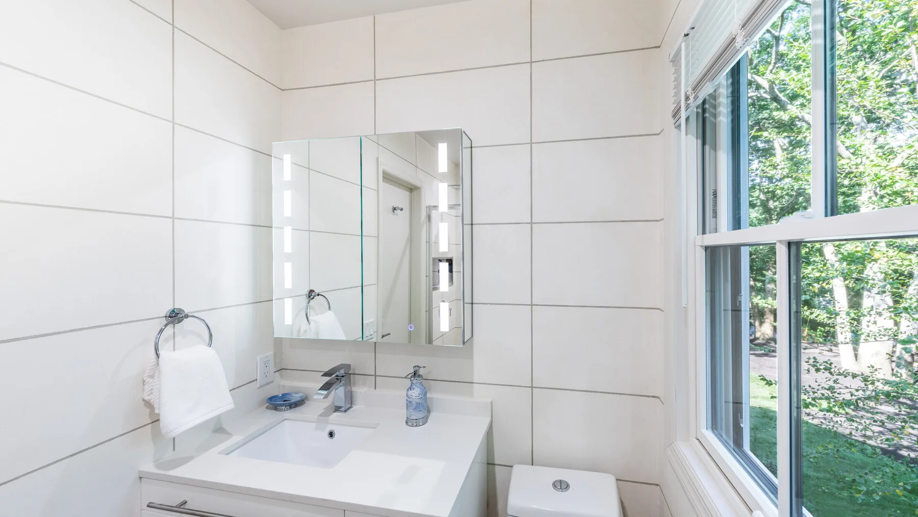 Un lavabo de baño luminoso con espejo y una ventana grande que da a la vegetación.
