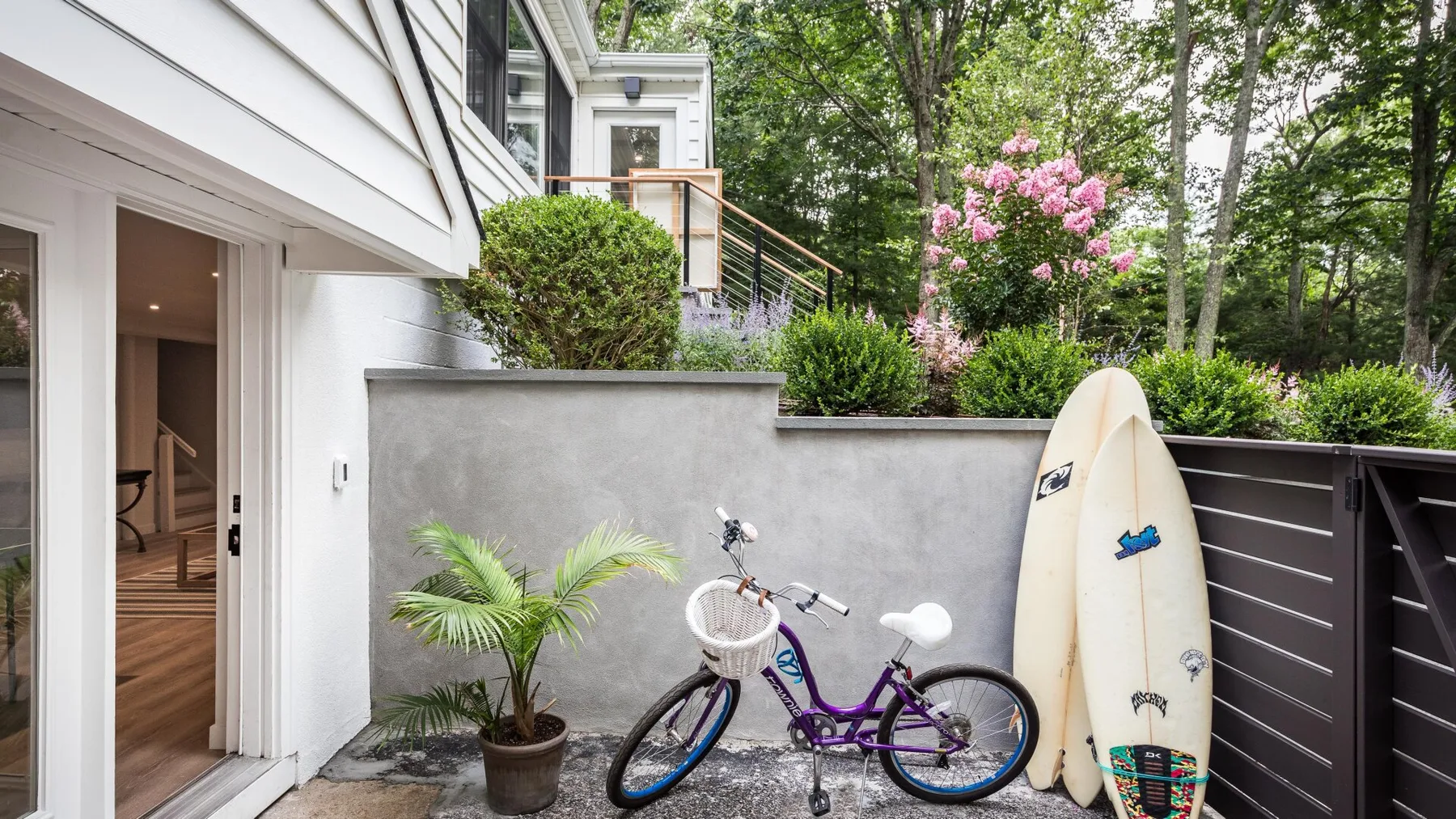 Un patio junto a una casa tiene una bicicleta y una tabla de surf junto a un muro bajo.