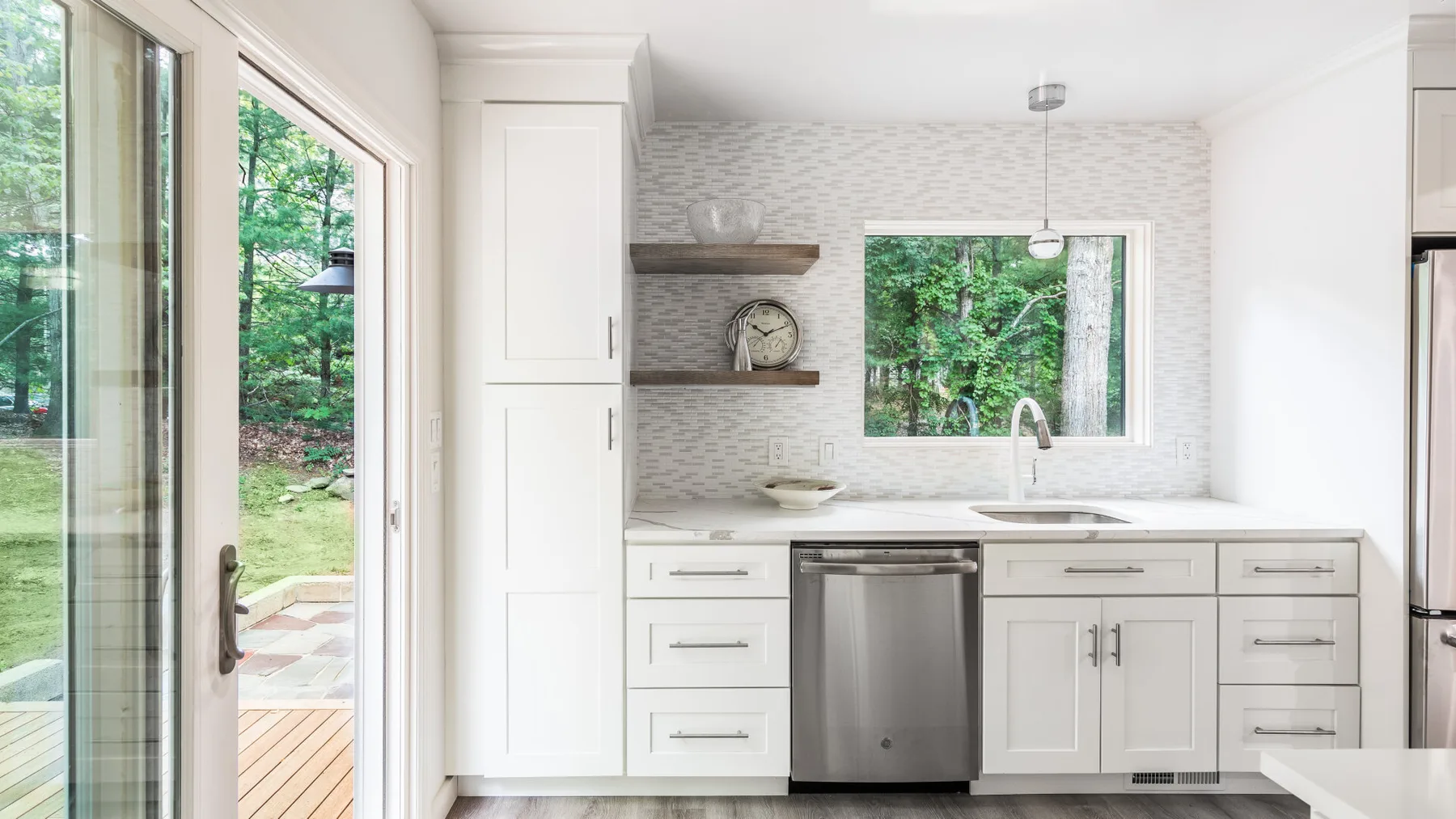 Una cocina luminosa con gabinetes blancos, lavavajillas de acero inoxidable y ventanas con vista a la vegetación.