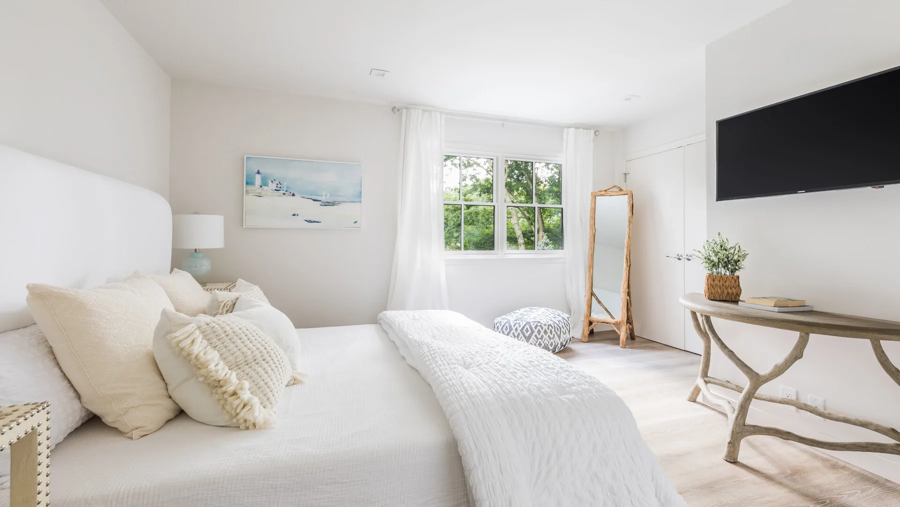 Un dormitorio luminoso con ropa de cama blanca, una ventana y un televisor en la pared.