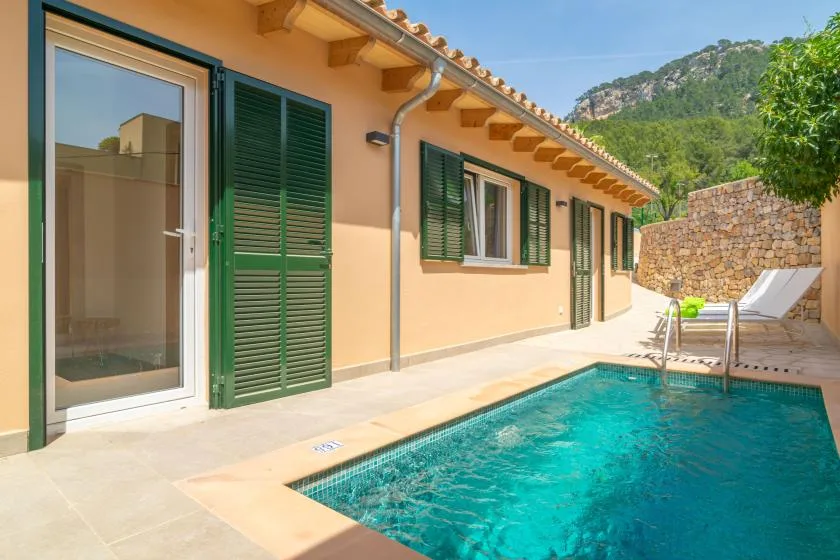 A swimming pool beside a house, with green shutters and a stone wall.