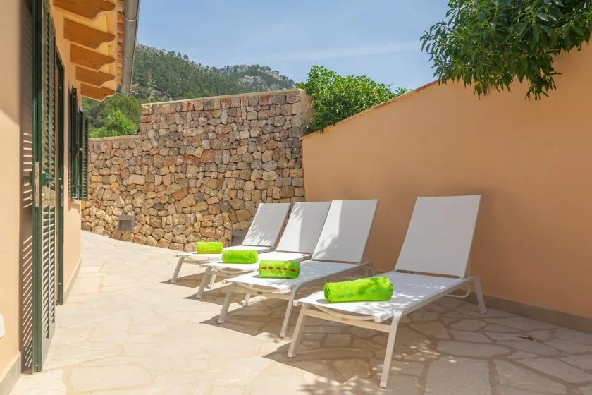 A sunlit outdoor terrace has white loungers beside a stone wall.