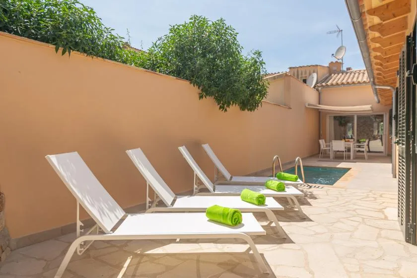 Un patio soleado junto a la piscina tiene tumbonas con toallas verdes.