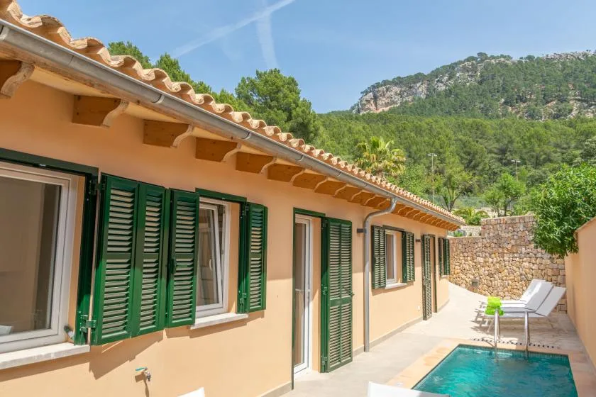 Un patio soleado con contraventanas verdes, una piscina y colinas boscosas al fondo.
