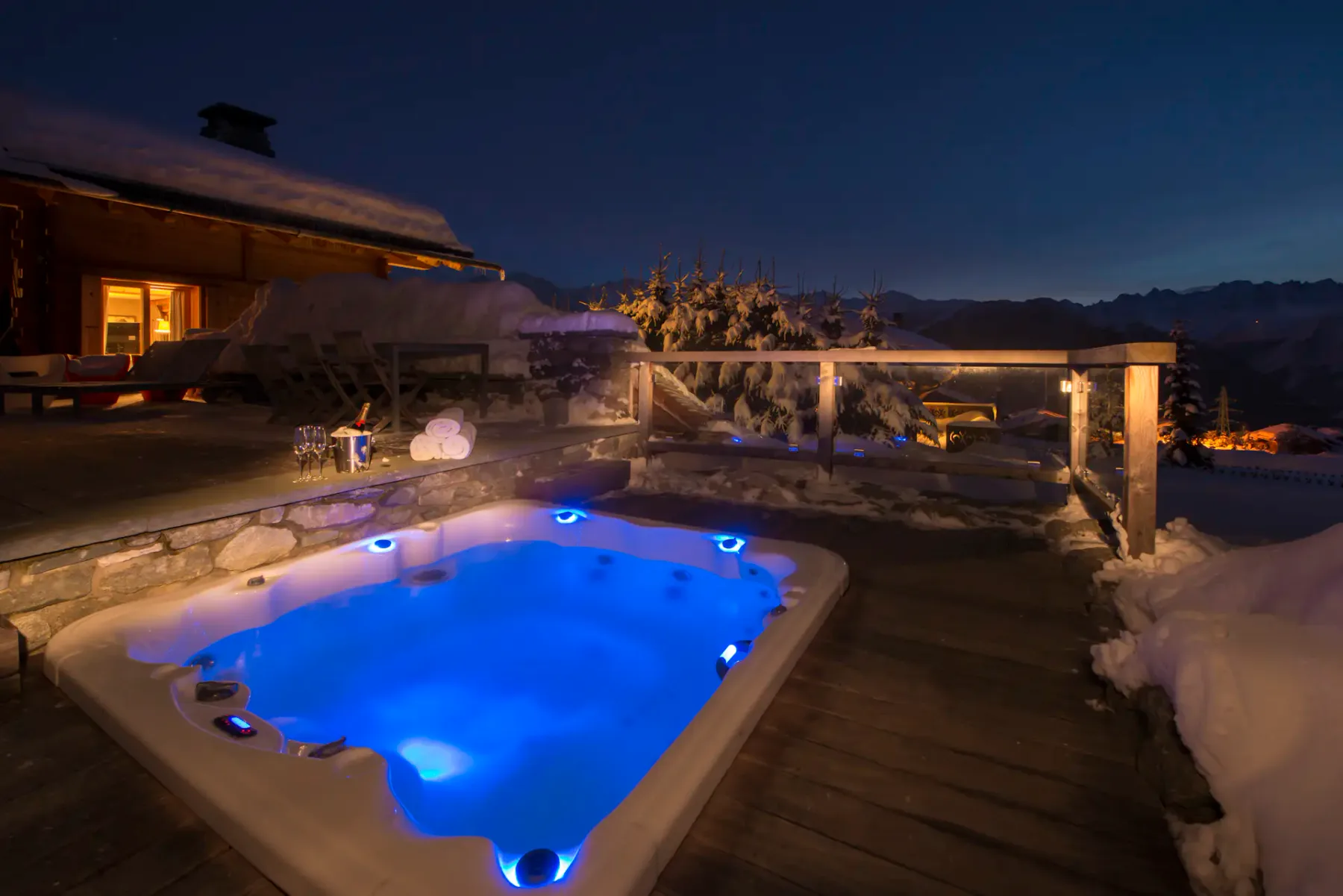 Bañera de hidromasaje al aire libre en una terraza de madera por la noche, rodeada de nieve, con una cabaña iluminada cerca y montañas visibles al fondo.