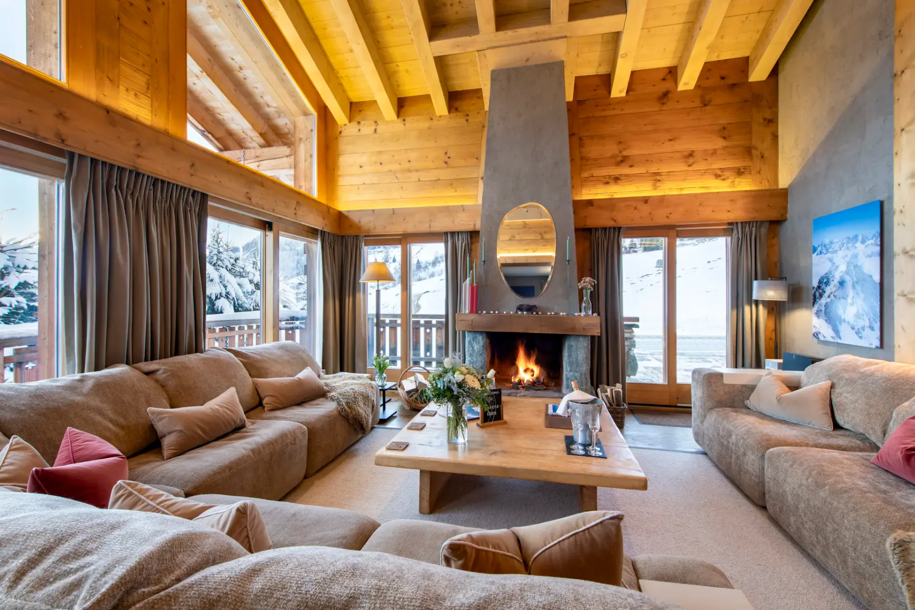 Acogedora sala de estar con grandes sofás, una mesa de centro, chimenea de piedra, techos de madera y grandes ventanales con vistas a un paisaje montañoso nevado.