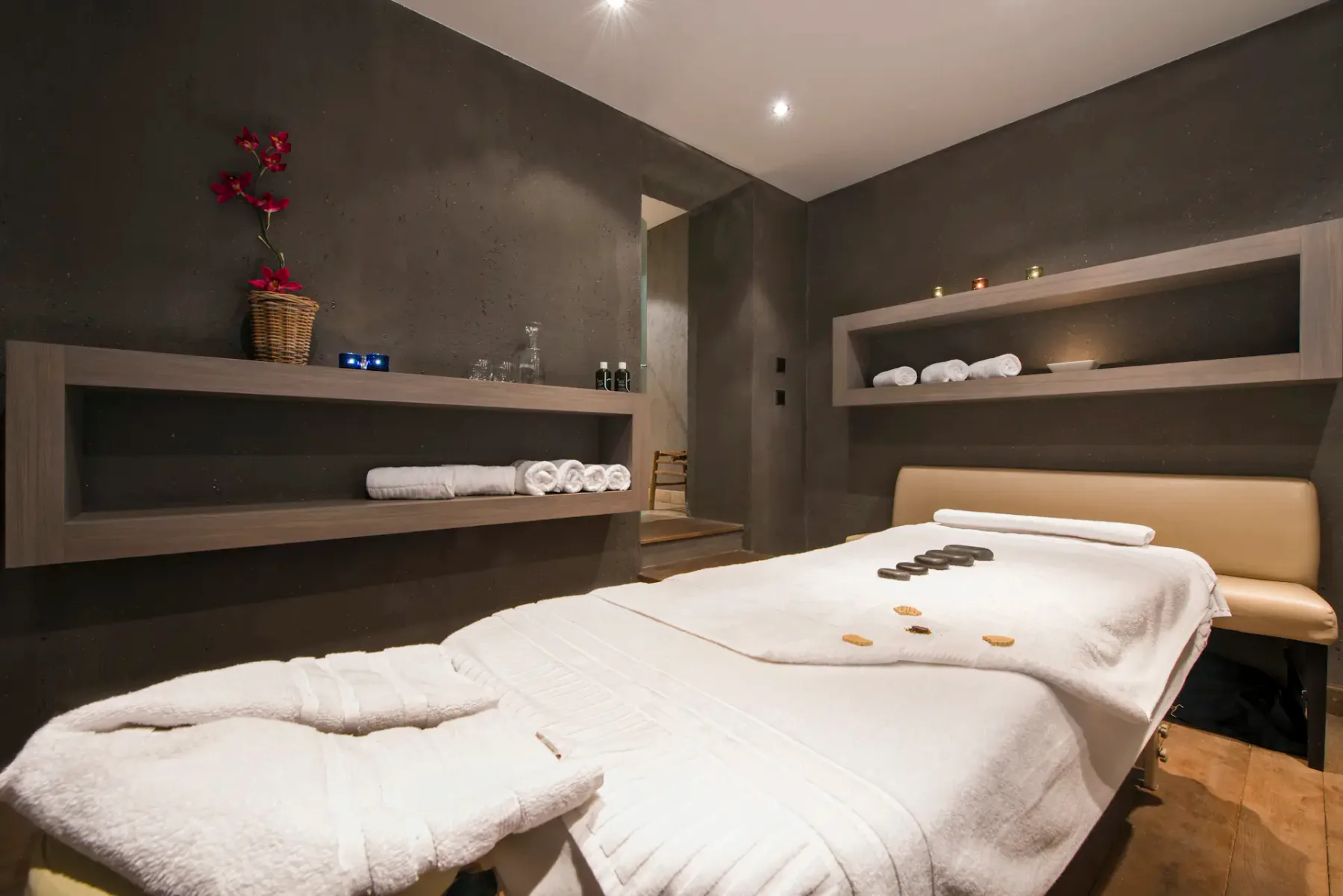 Moderna sala de tratamientos de spa con una camilla de masaje cubierta con toallas blancas, piedras calientes dispuestas encima y estantes con toallas enrolladas y objetos decorativos.