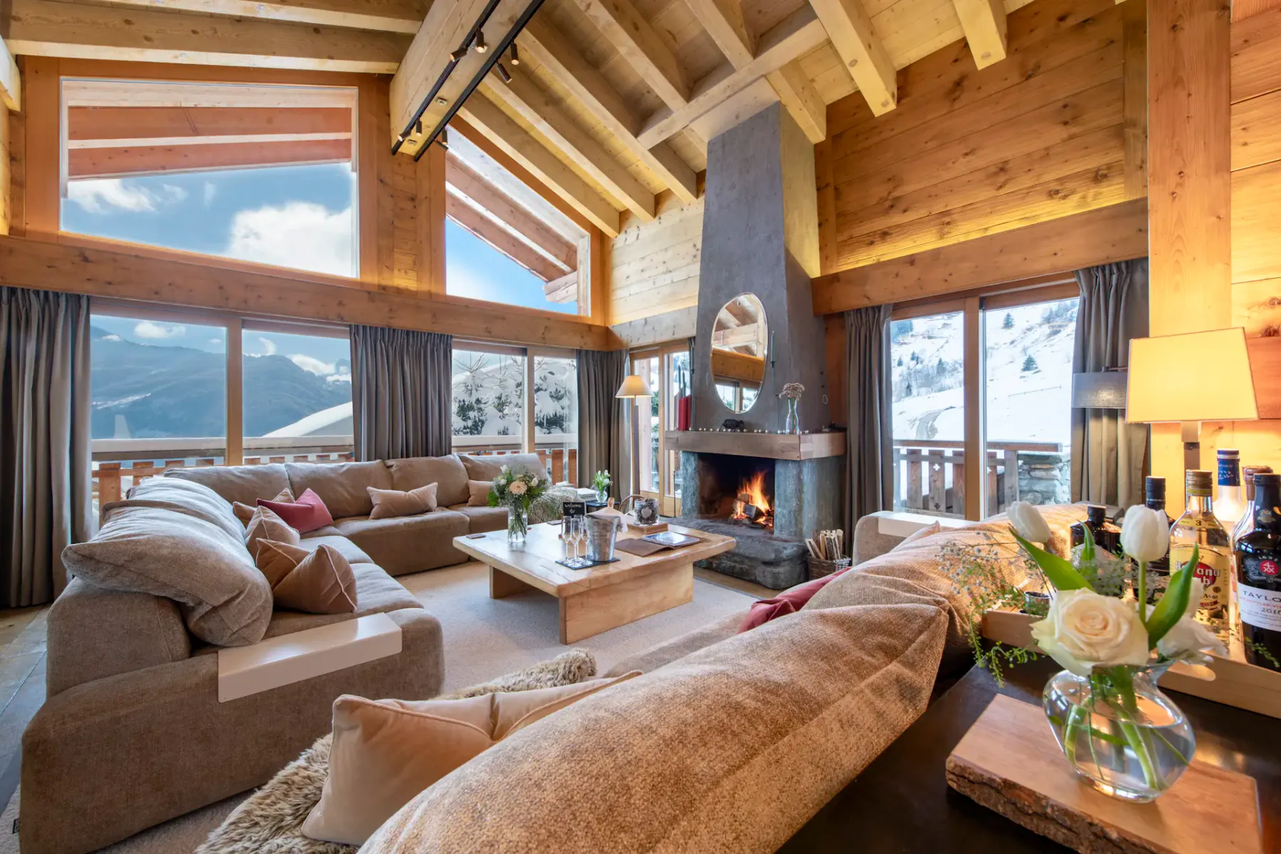 Acogedora sala de estar con grandes sofás, una mesa de centro, chimenea de piedra, techos de madera y grandes ventanales con vistas a un paisaje montañoso nevado.