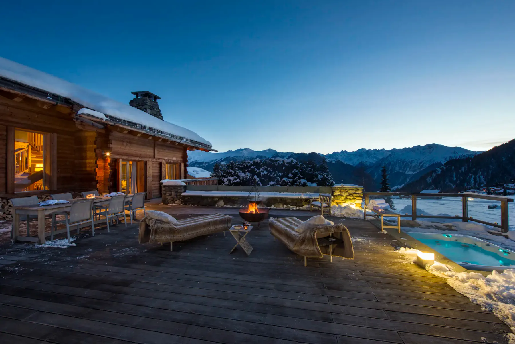 Bañera de hidromasaje al aire libre en una terraza de madera por la noche, rodeada de nieve, con una cabaña iluminada cerca y montañas visibles al fondo.