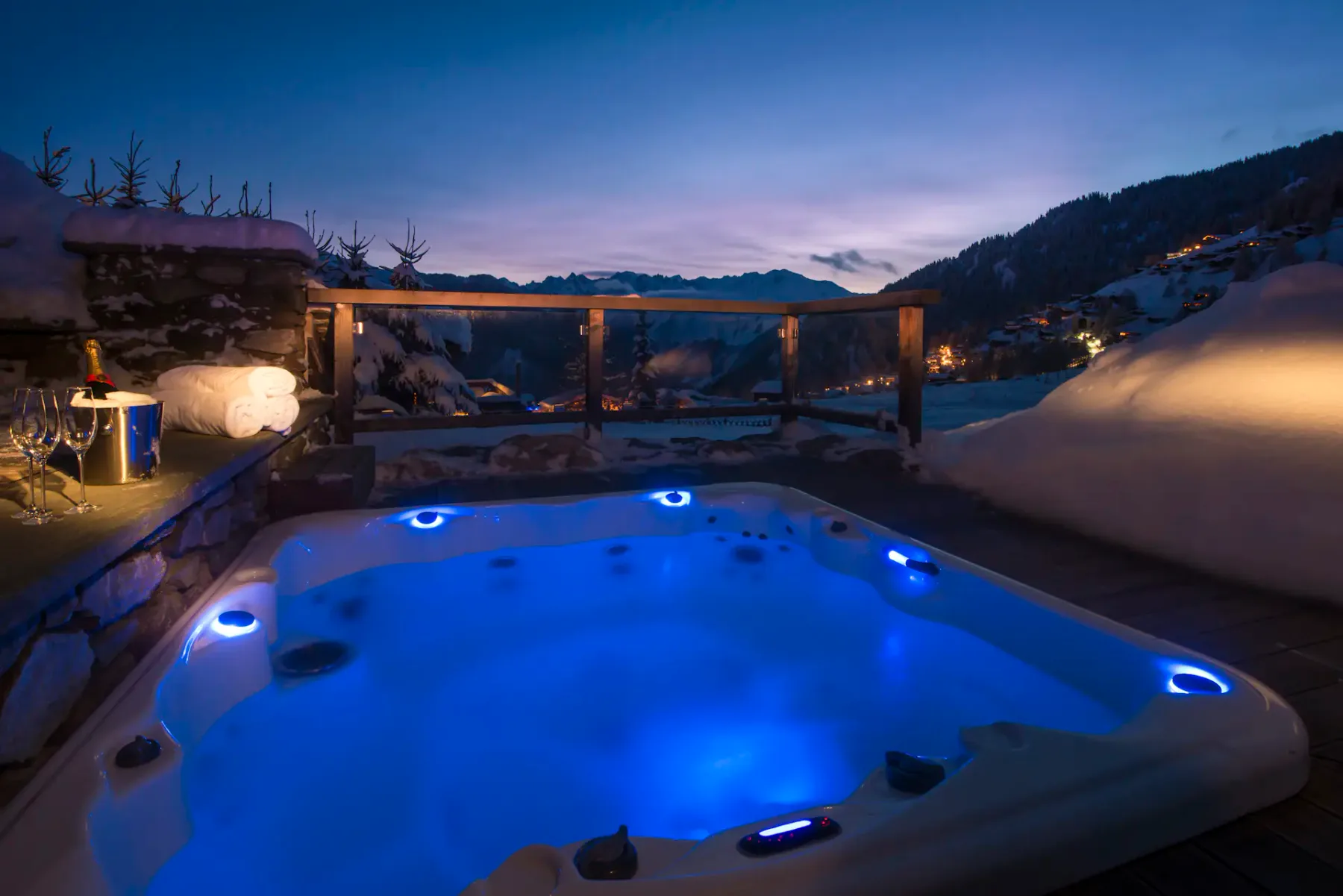 Bañera de hidromasaje al aire libre en una terraza de madera por la noche, rodeada de nieve, con una cabaña iluminada cerca y montañas visibles al fondo.