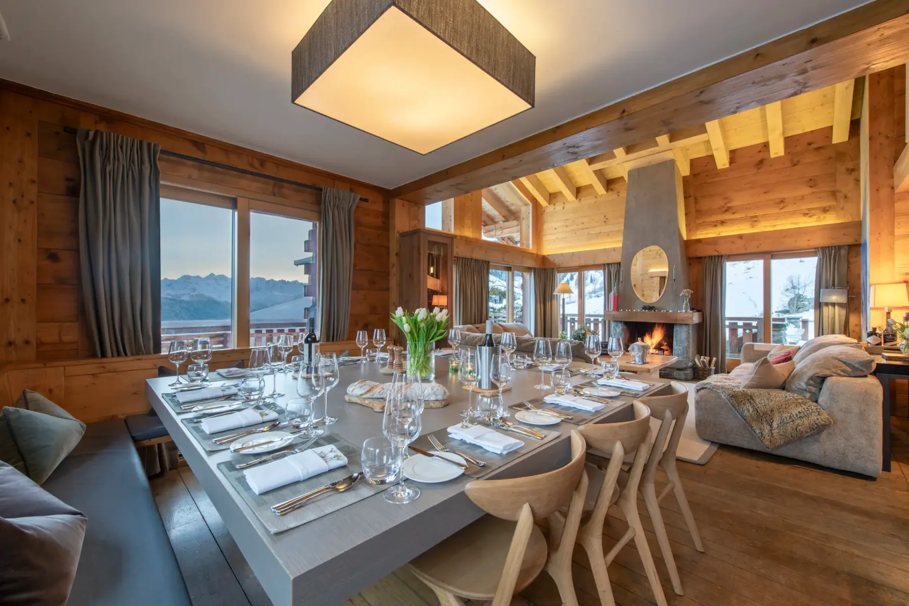Comedor de madera con una mesa puesta para diez personas, una lámpara moderna, chimenea, una acogedora zona de estar, grandes ventanales y vistas a las montañas.