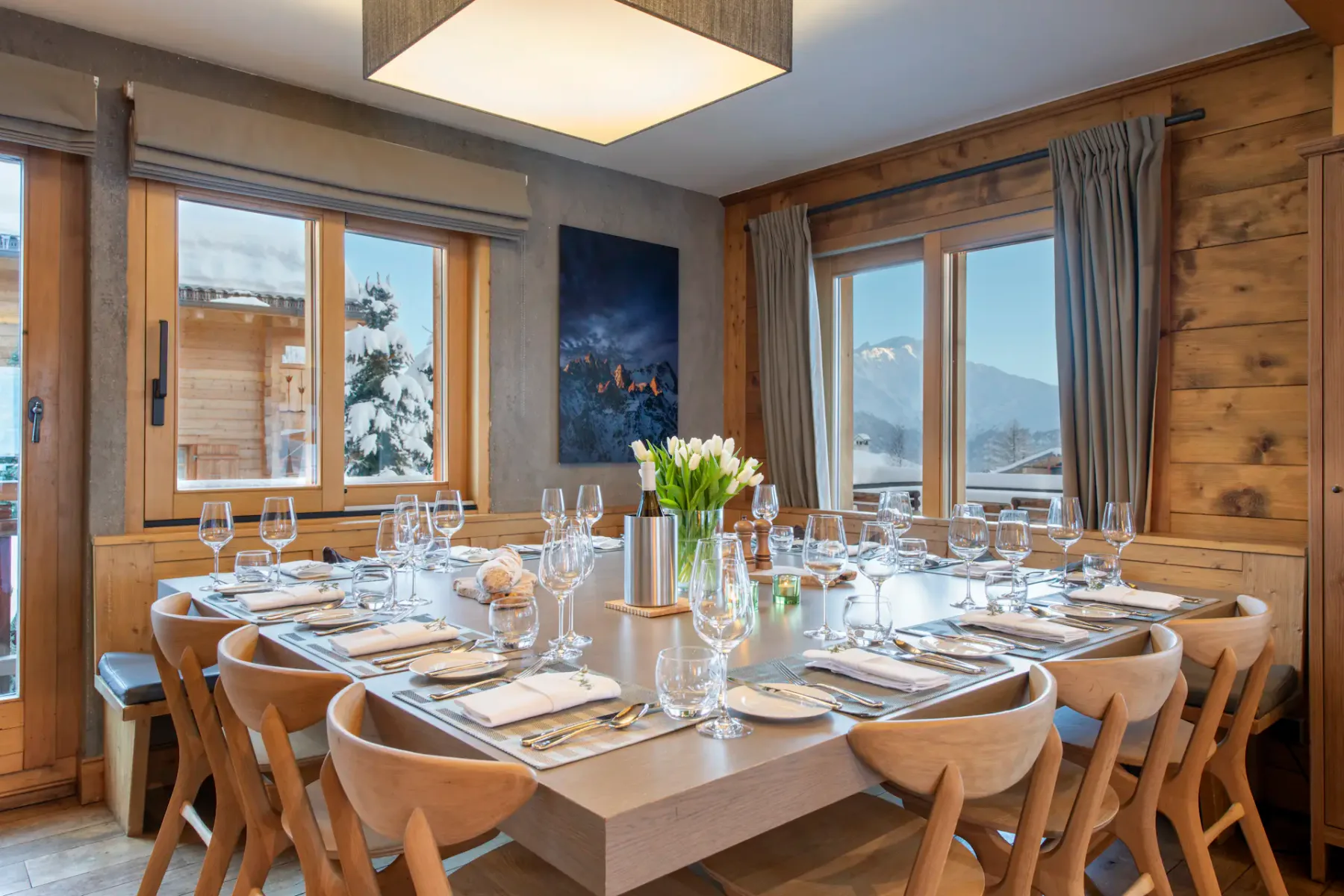Comedor de madera con una mesa puesta para diez personas, una lámpara moderna, chimenea, una acogedora zona de estar, grandes ventanales y vistas a las montañas.