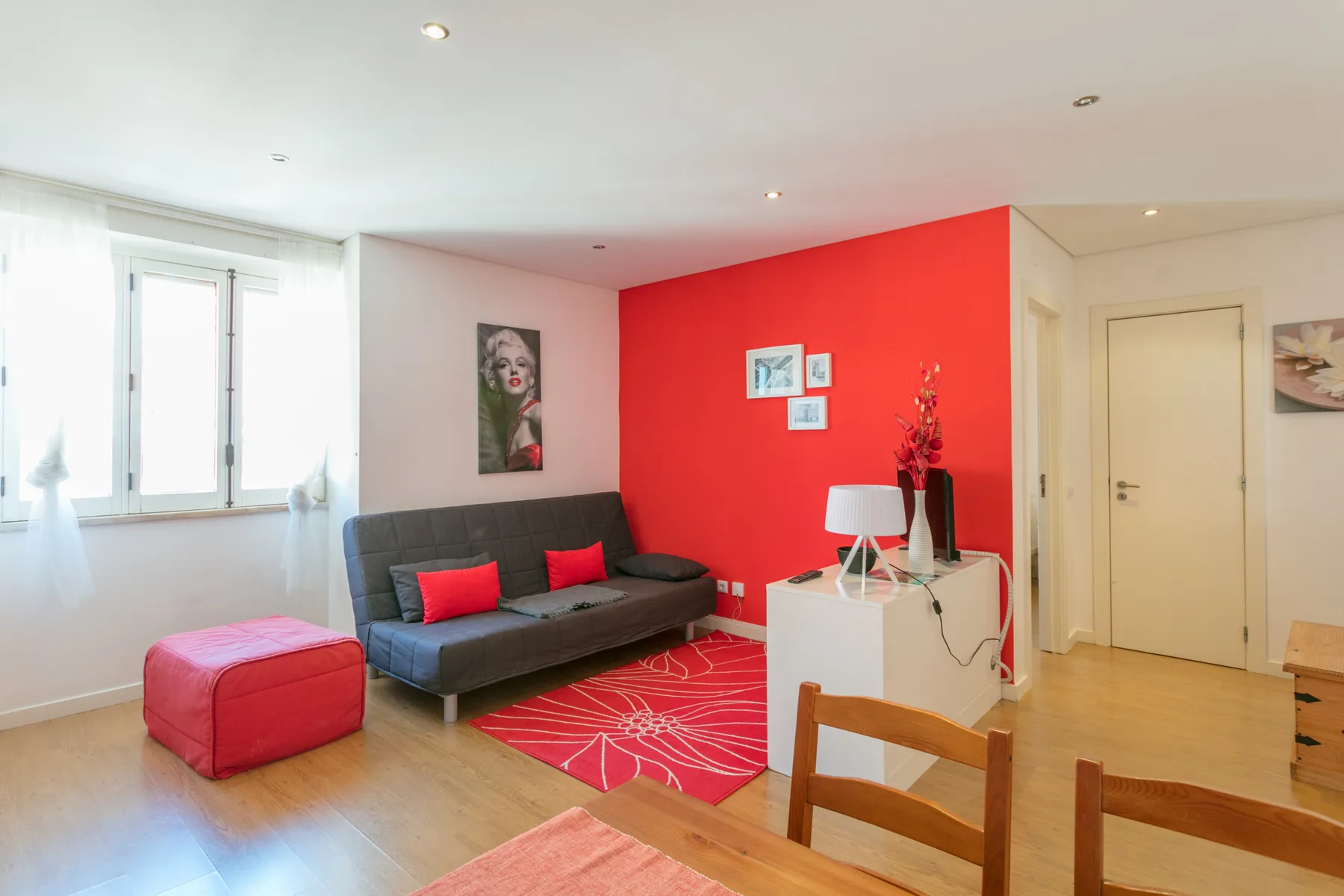 Una sala colorida tiene un sofá gris, una alfombra roja y una ventana luminosa.