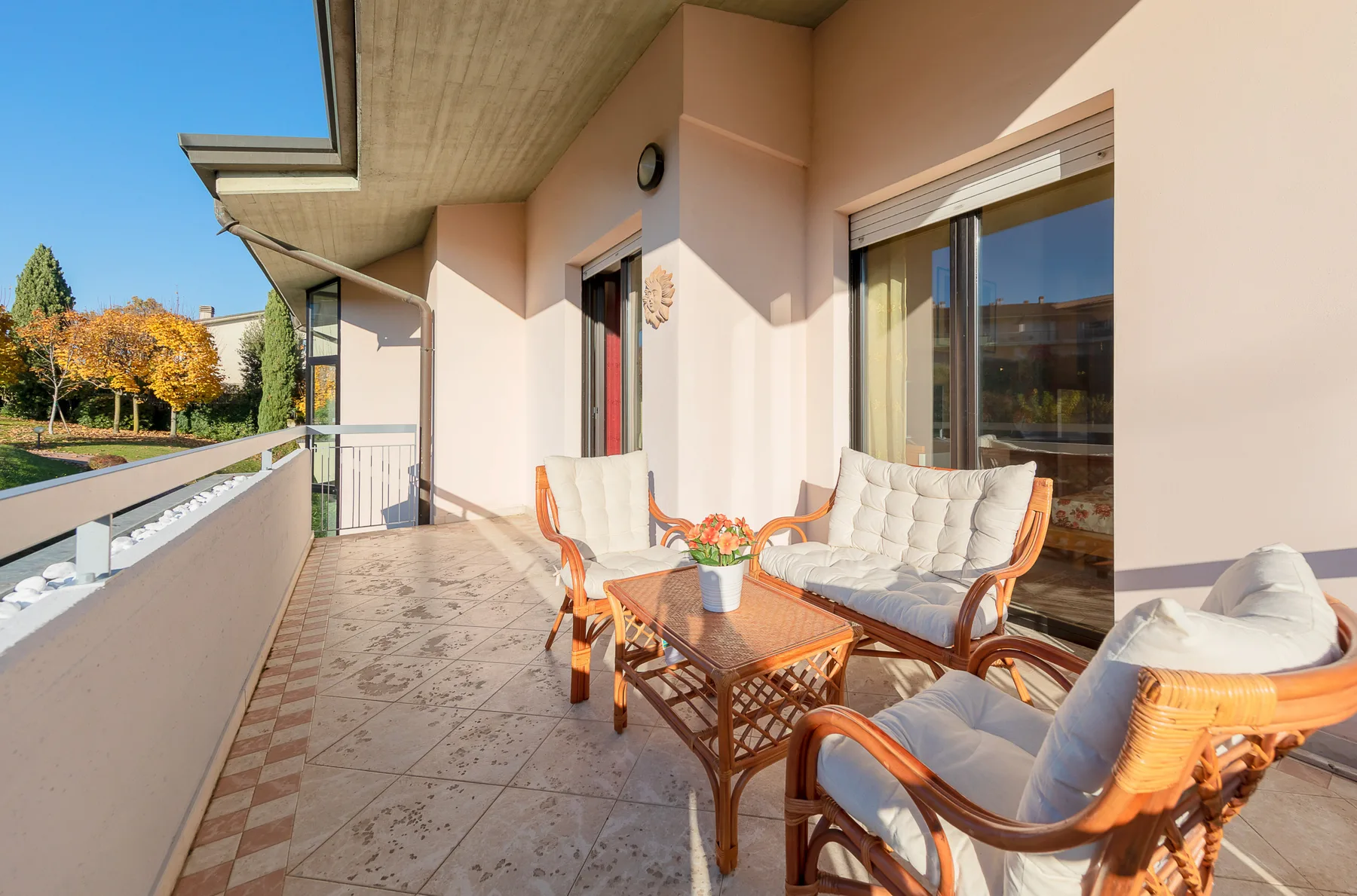Una terraza soleada con asientos de mimbre y una mesa baja junto a puertas de vidrio.