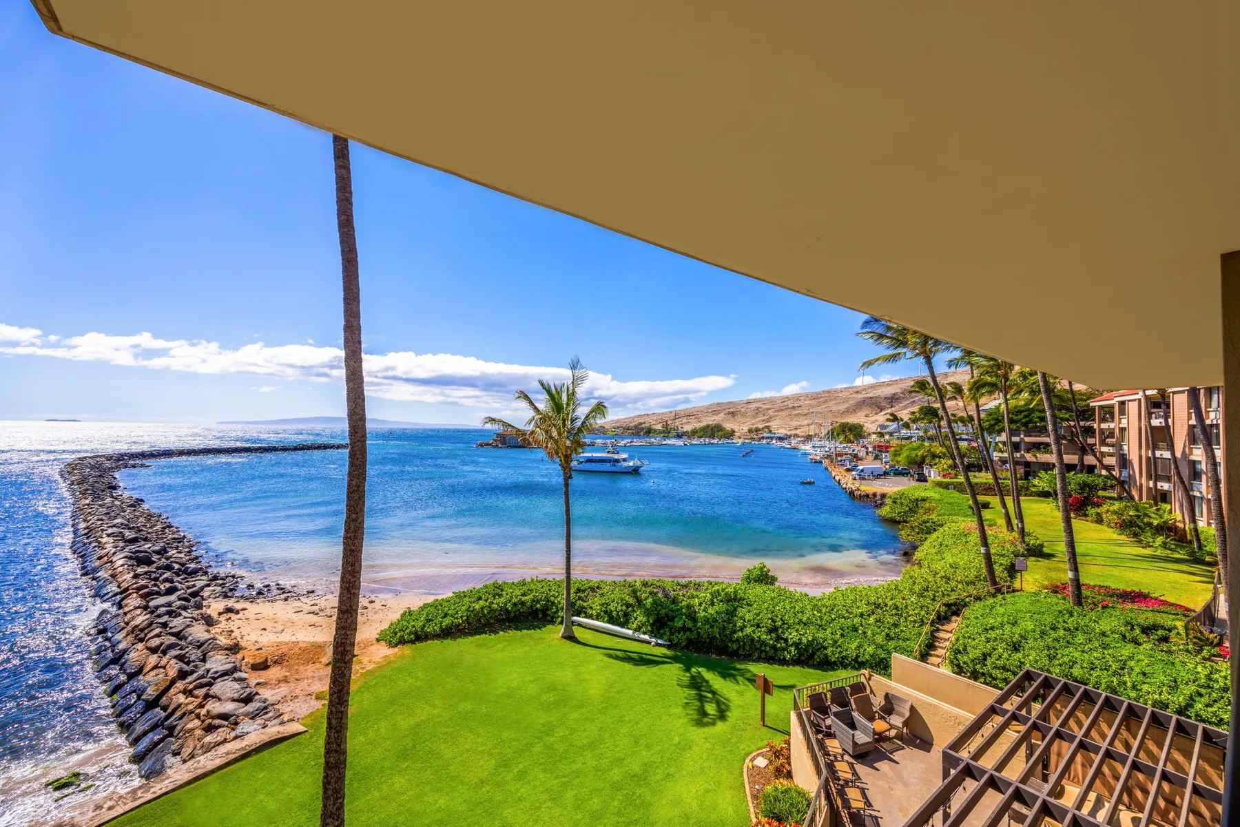 A coastal resort lawn overlooks a blue bay, with palm trees and a rocky shoreline.