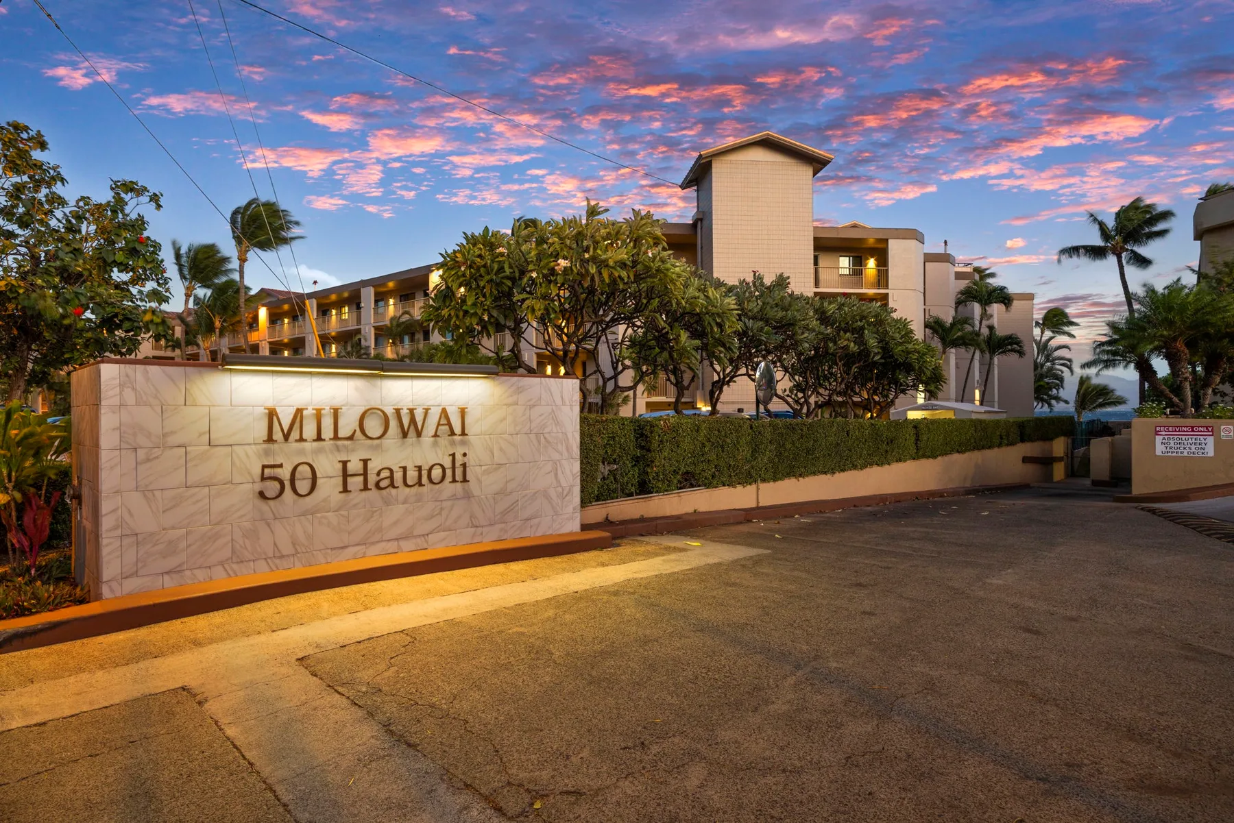 A landscaped residential building stands behind a stone entrance sign at sunset.