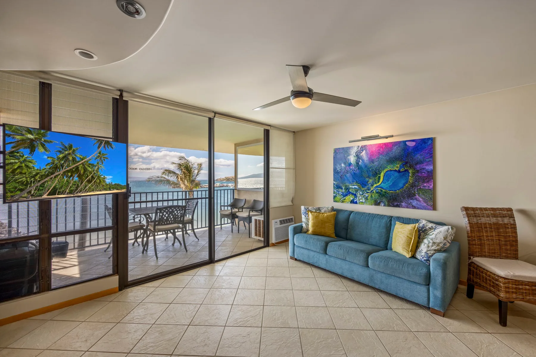 A living room opens onto a balcony, with blue seating and a colorful wall painting.