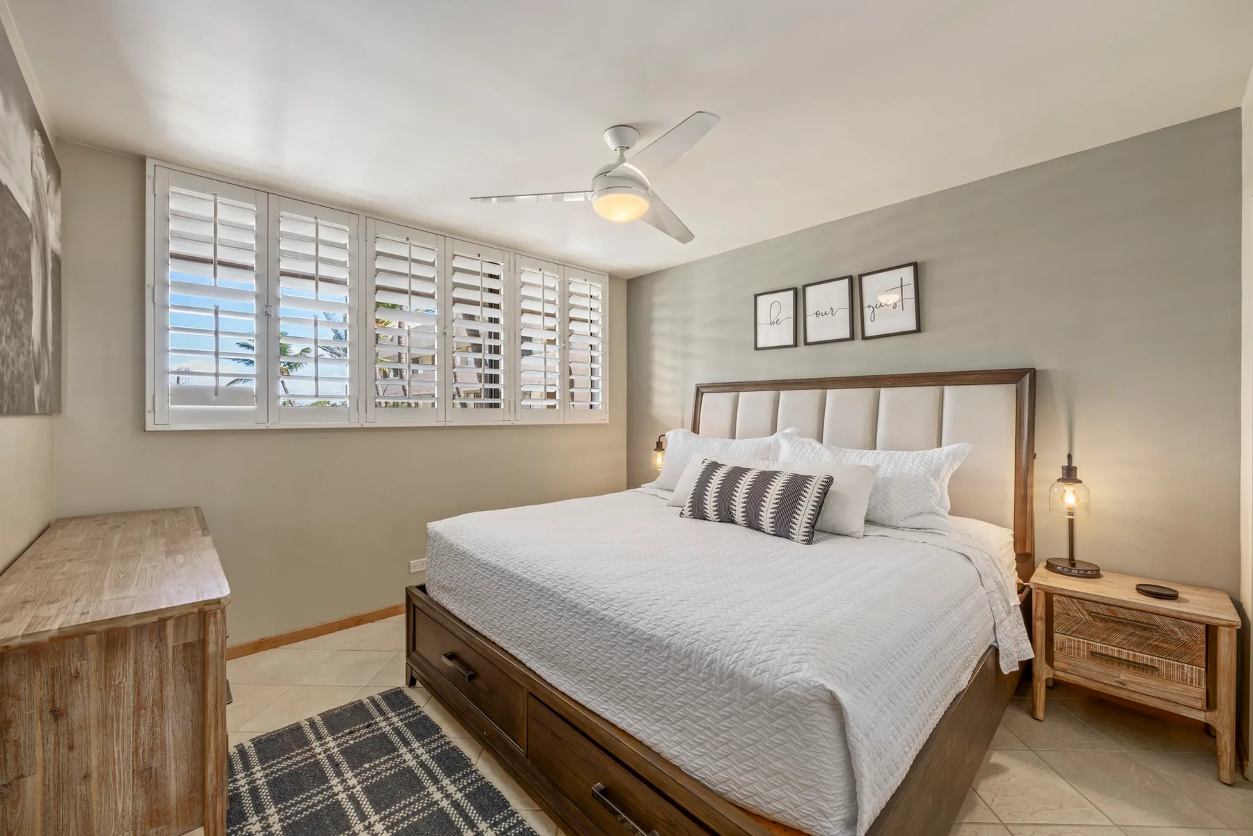A bedroom with a bed beneath shuttered windows and a ceiling fan.
