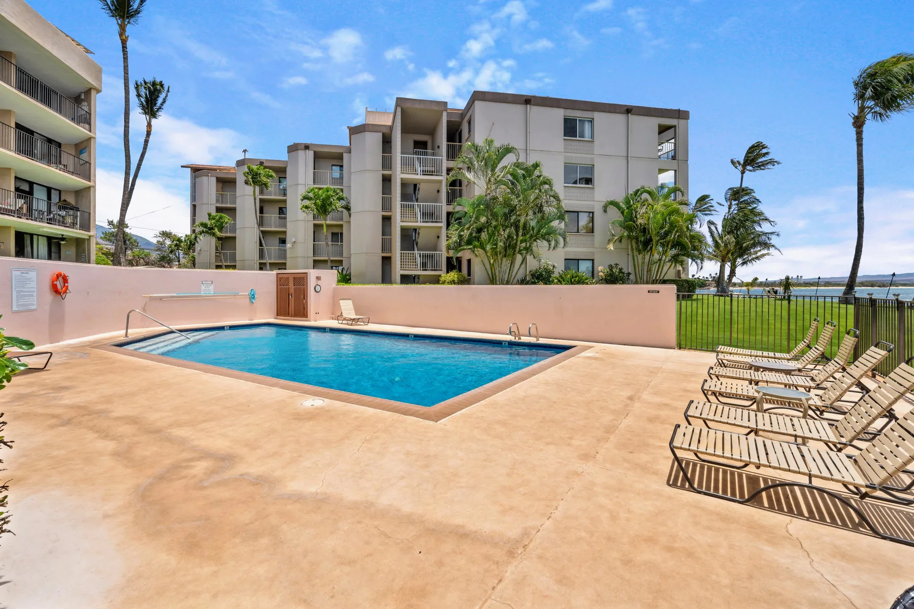 An outdoor swimming pool sits beside apartment buildings, with lounge chairs along the deck.