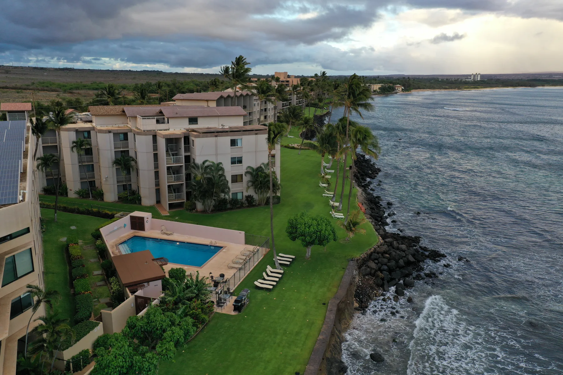 A coastal resort complex borders the rocky shoreline, with a pool and palm-lined lawn.
