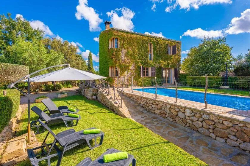 Un patio ajardinado junto a una piscina, con asientos reclinables y una casa cubierta de enredaderas.