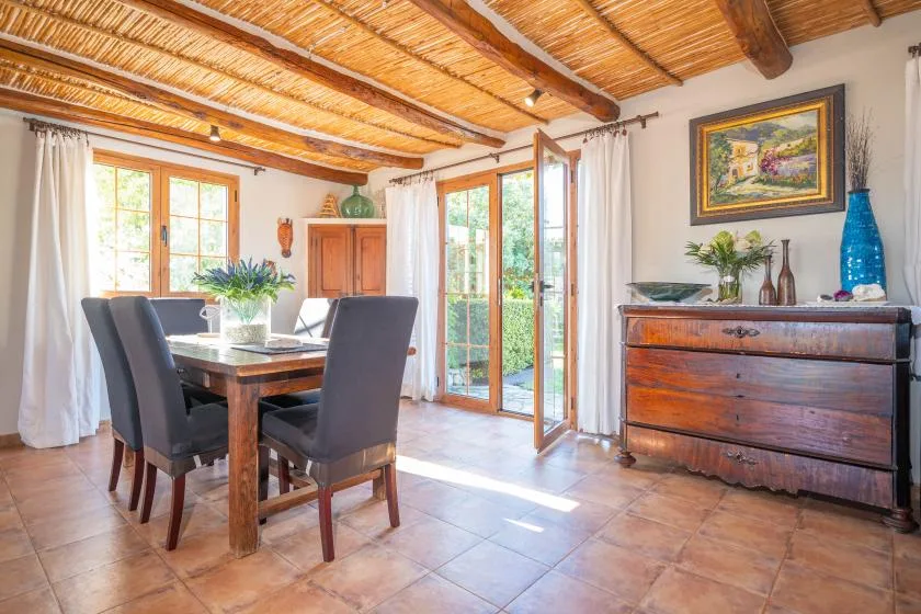 Un comedor luminoso tiene una mesa de madera, asientos oscuros y grandes ventanas.