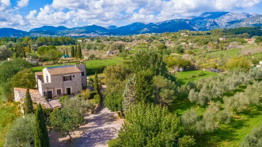 Una casa de piedra se alza entre campos verdes, con montañas a lo lejos.