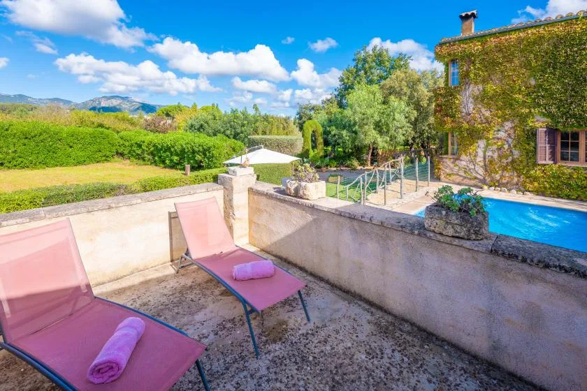 Un patio soleado da a una piscina, con asientos reclinables rosados y vegetación.