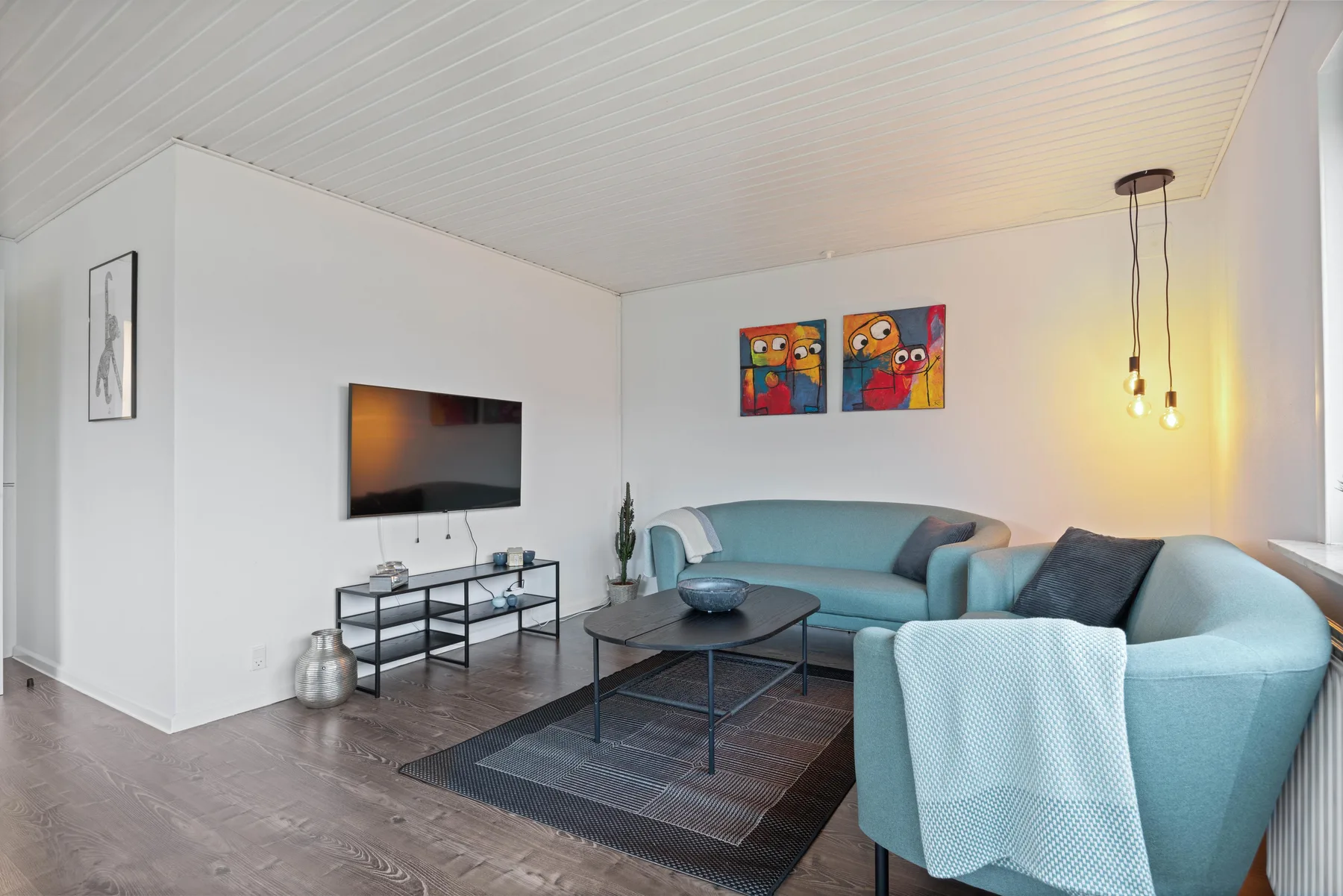 Sala de estar con asientos azules, un televisor y cuadros coloridos.