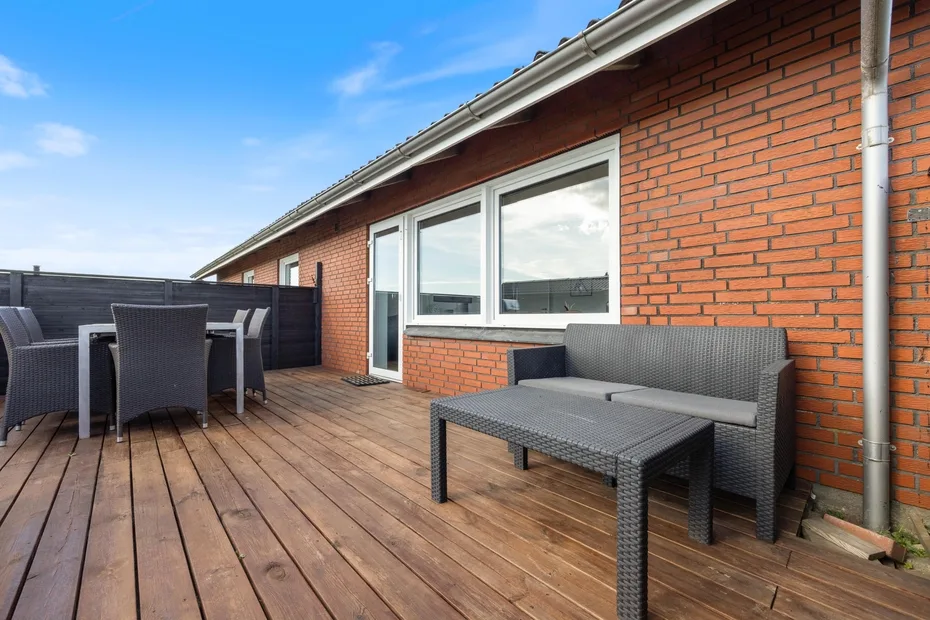 Una terraza de madera junto a un edificio de ladrillo tiene asientos al aire libre y ventanas grandes.