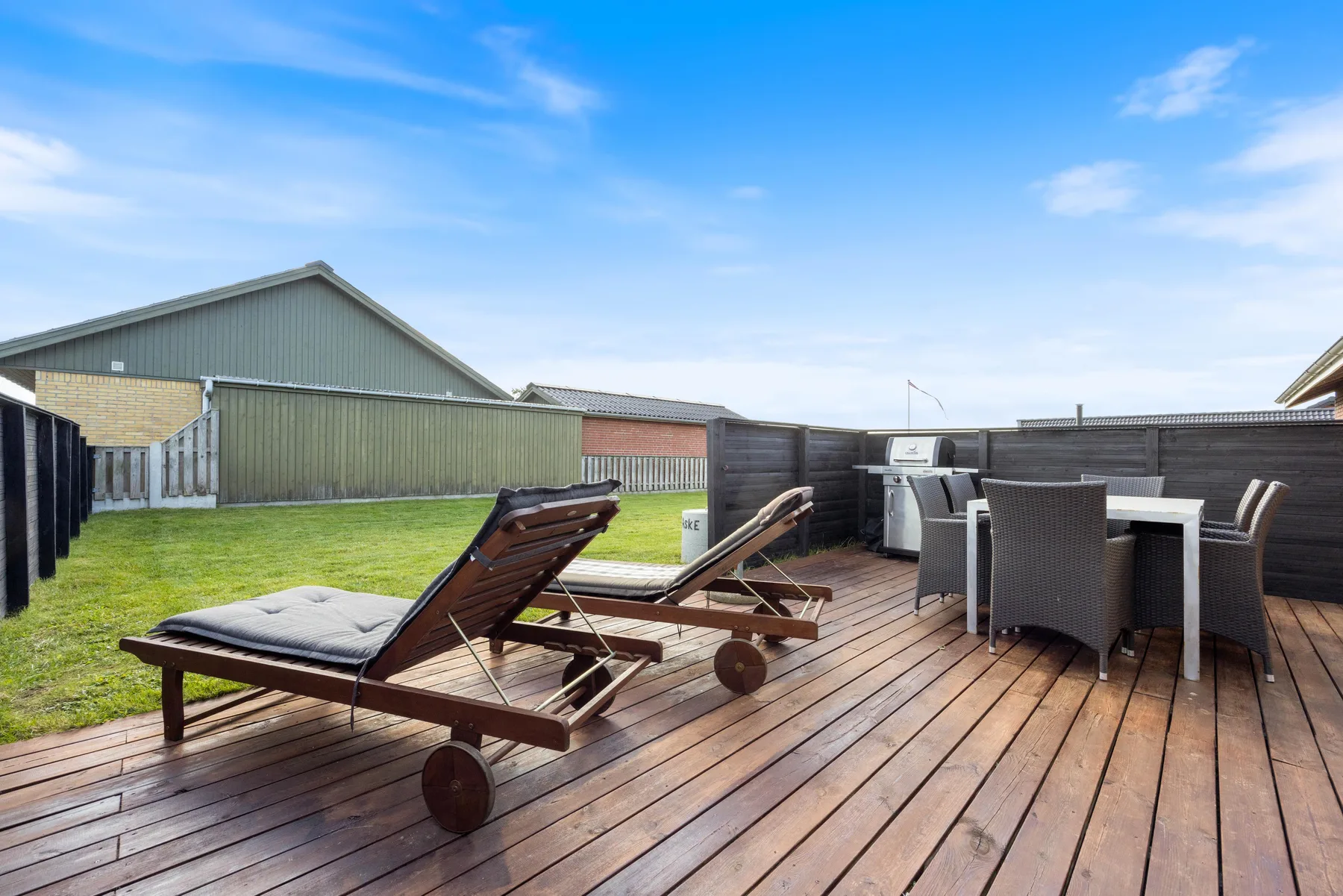 Una terraza de madera tiene tumbonas reclinables y asientos de comedor junto a un jardín con césped.