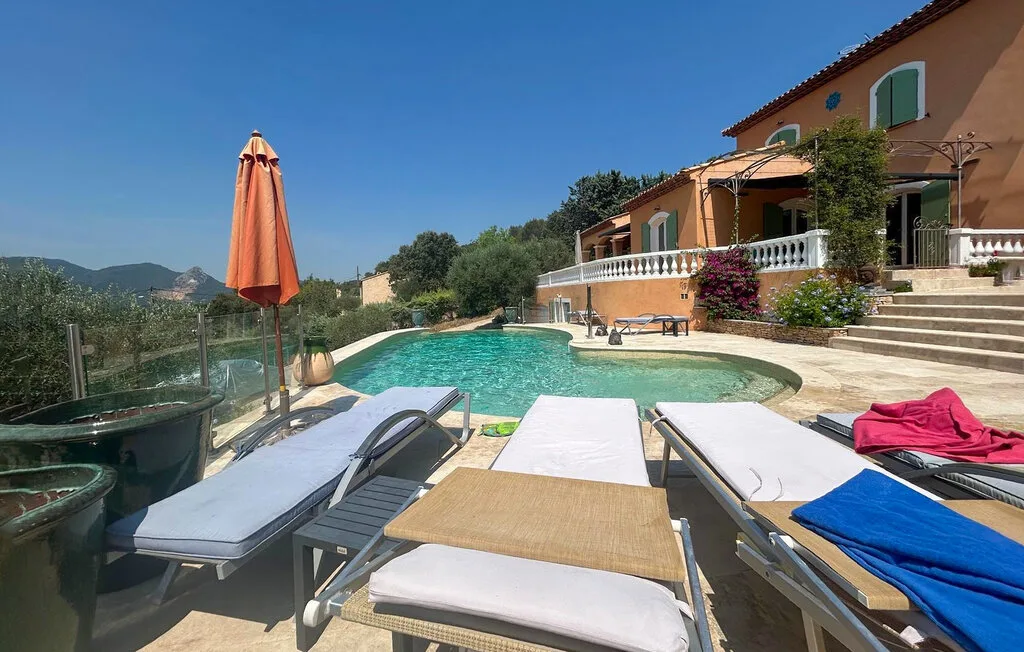 Una piscina al aire libre junto a una casa, con asientos reclinables y una sombrilla cerrada.