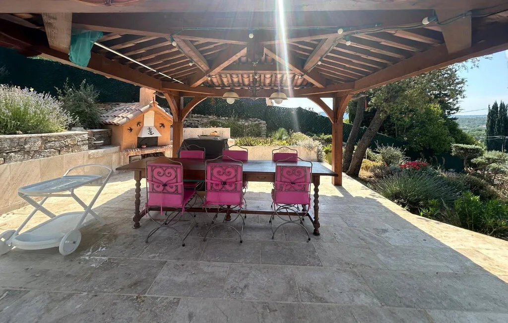 Una zona de comedor exterior cubierta con mesa y asientos rosas da a un jardín.
