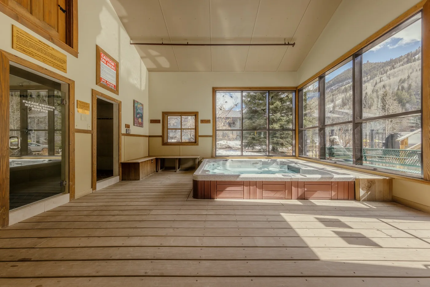 Una zona interior soleada con jacuzzi y grandes ventanas que dan a una ladera arbolada.
