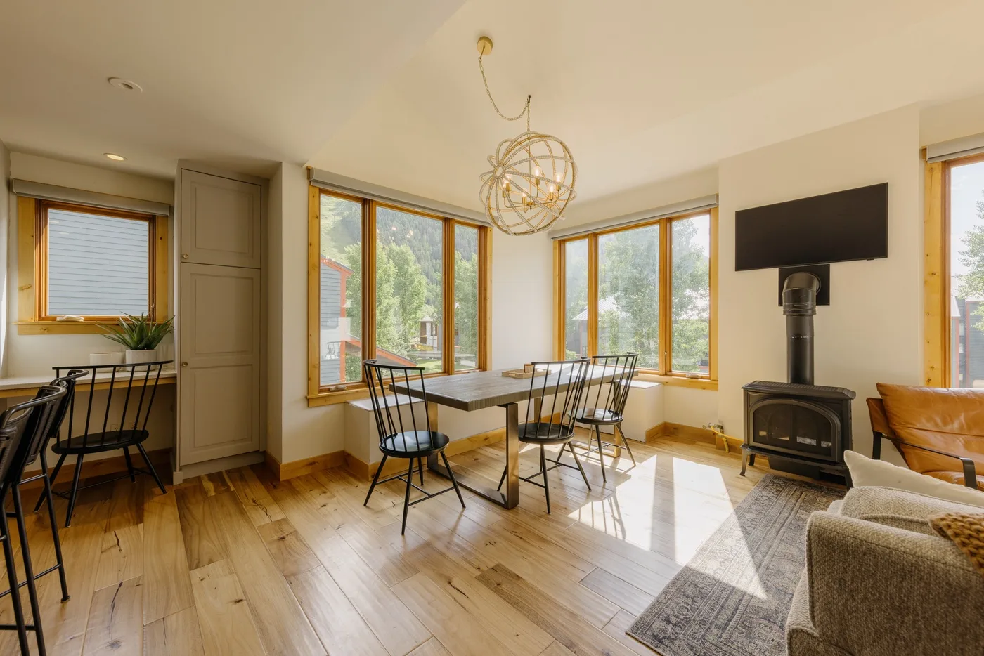 Una luminosa sala de estar y comedor tiene grandes ventanas, una mesa de comedor y una estufa de leña.