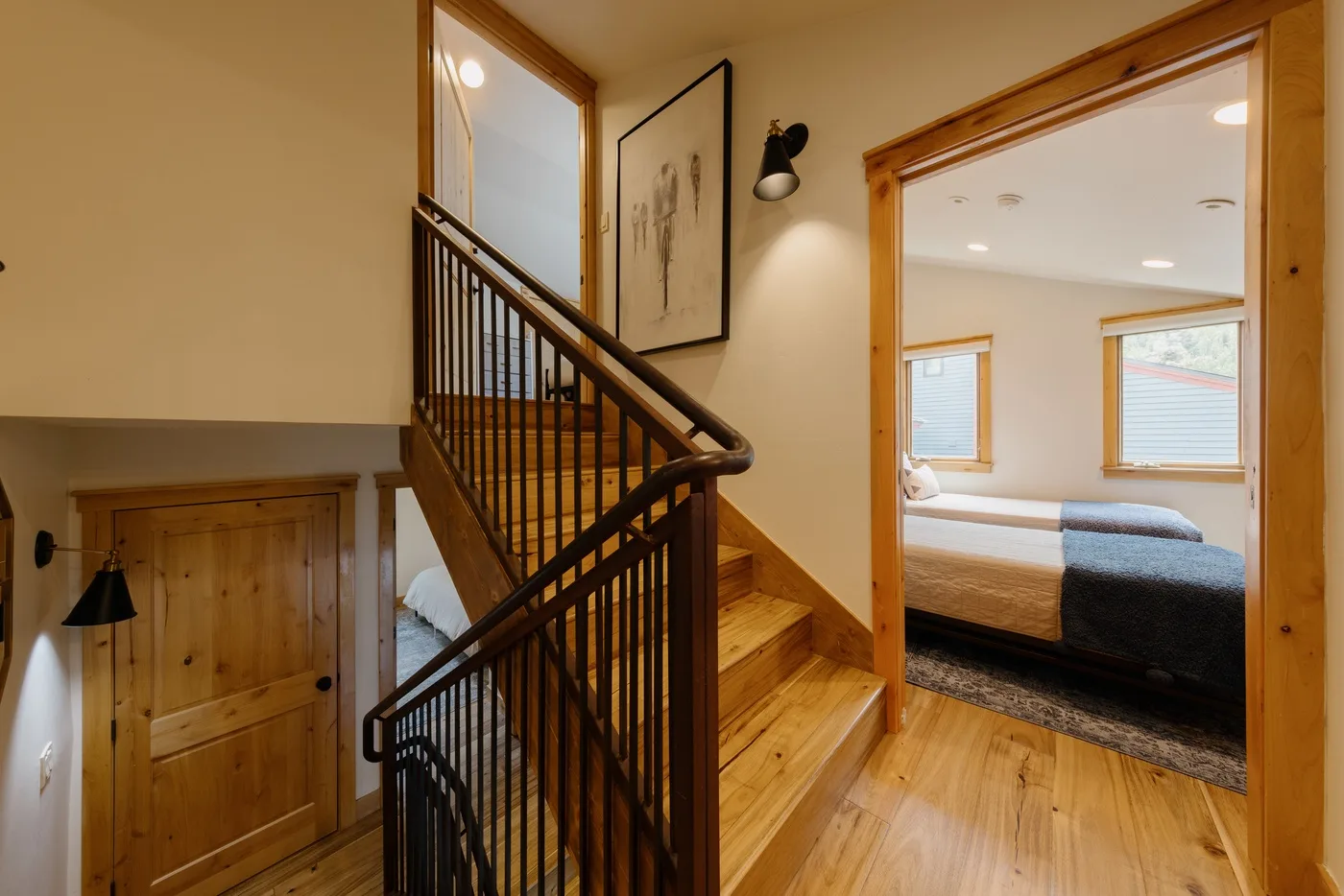 Una escalera de madera y un pasillo dan a un dormitorio con una cama y ventanas.