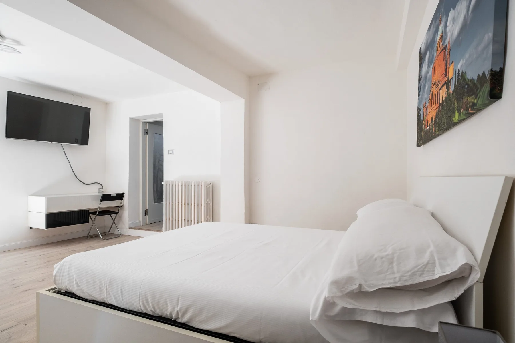 Un dormitorio luminoso con una cama blanca, un televisor en la pared y un escritorio.